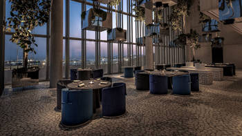 A space with a mosaic floor, round tables and a glass window facing outside, decorated with square lamps hanging together with tree branches.